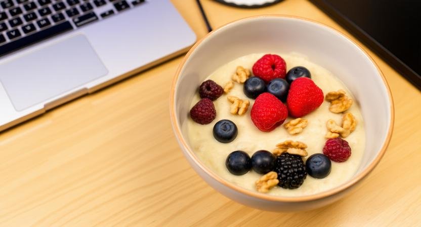 A nutritious bowl of oatmeal topped with fresh berries and walnuts placed on an office desk next to a laptop.